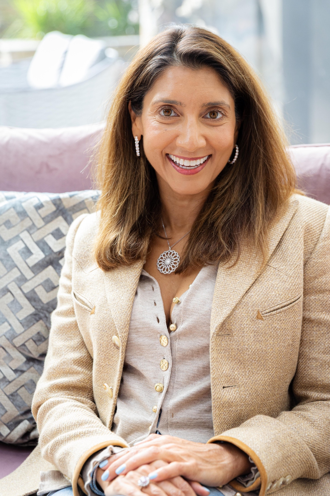 Aliya Kanji seated on a soft mauve sofa wearing a camel tweed blazer, smiling warmly.